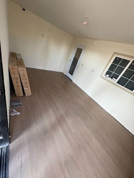 Room with light brown hardwood floors, beige walls, and boxes; a door and window are visible.