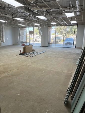 Empty commercial space, concrete floor, large windows, exposed ceiling, construction materials, and a vehicle visible outside.