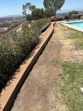 A long, low retaining wall separates dry dirt from grassy area beside a pool, overlooking a distant valley.