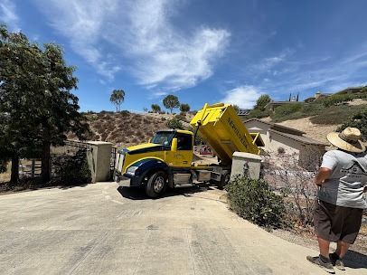 Yellow dump truck backed into driveway, dumping debris; person watches. Outdoors, sunny.