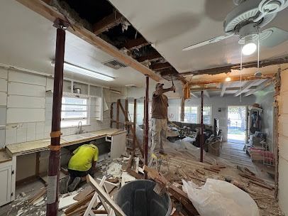 Men demolish a kitchen. Debris litters the floor. One works on a beam, while another works below.