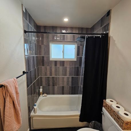 Bathroom with gray tile, white tub, black shower curtain, and a small window. Pink towel hanging.