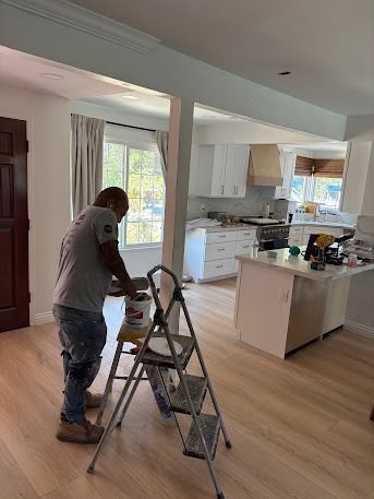 A man painting a wall in a home. White walls, light wood floors, and a kitchen in the background.