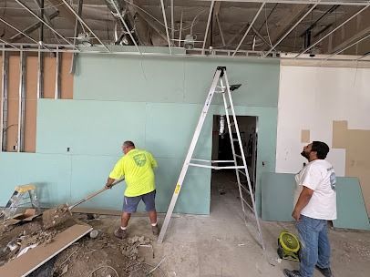 Two workers inside, installing drywall. One sweeps, the other watches. Interior construction site.