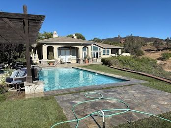 A swimming pool and house in bright sunlight. Green grass, a pergola, and a hill are visible.