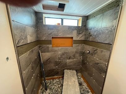 Shower stall with gray tiles, built-in niche, small window, and wood and pebble floor.