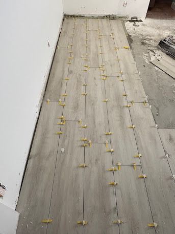 Gray wood-look floor tiles being installed, with yellow spacers visible, inside a room with white walls.