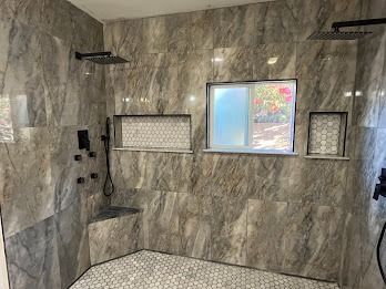 Luxurious shower with marble-like tile, two showerheads, window, and built-in shelves, in a neutral-toned bathroom.