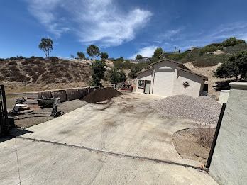 A driveway under construction with piles of gravel, dirt, and a building, against a hillside and a blue sky.