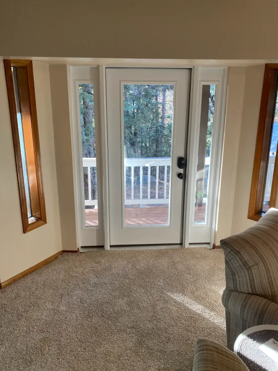 A white door with sidelights leads to a deck, flanked by wood-trimmed windows in a carpeted room.