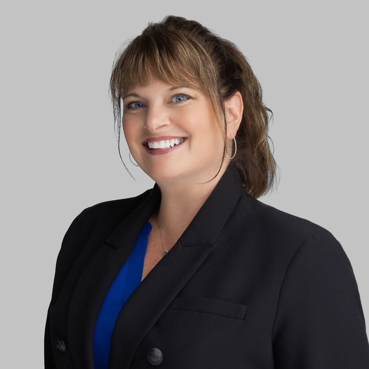 Woman with dark hair smiles, wearing a dark blazer, against a light gray background.
