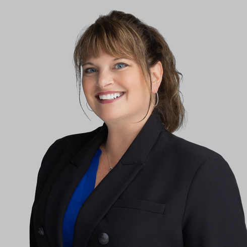 Woman with dark hair smiles, wearing a dark blazer, against a light gray background.