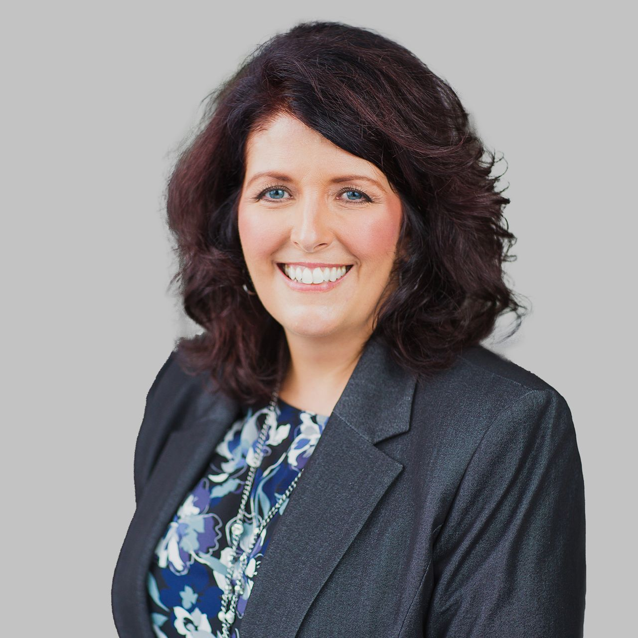 Woman with dark hair smiles, wearing a dark blazer, against a light gray background.