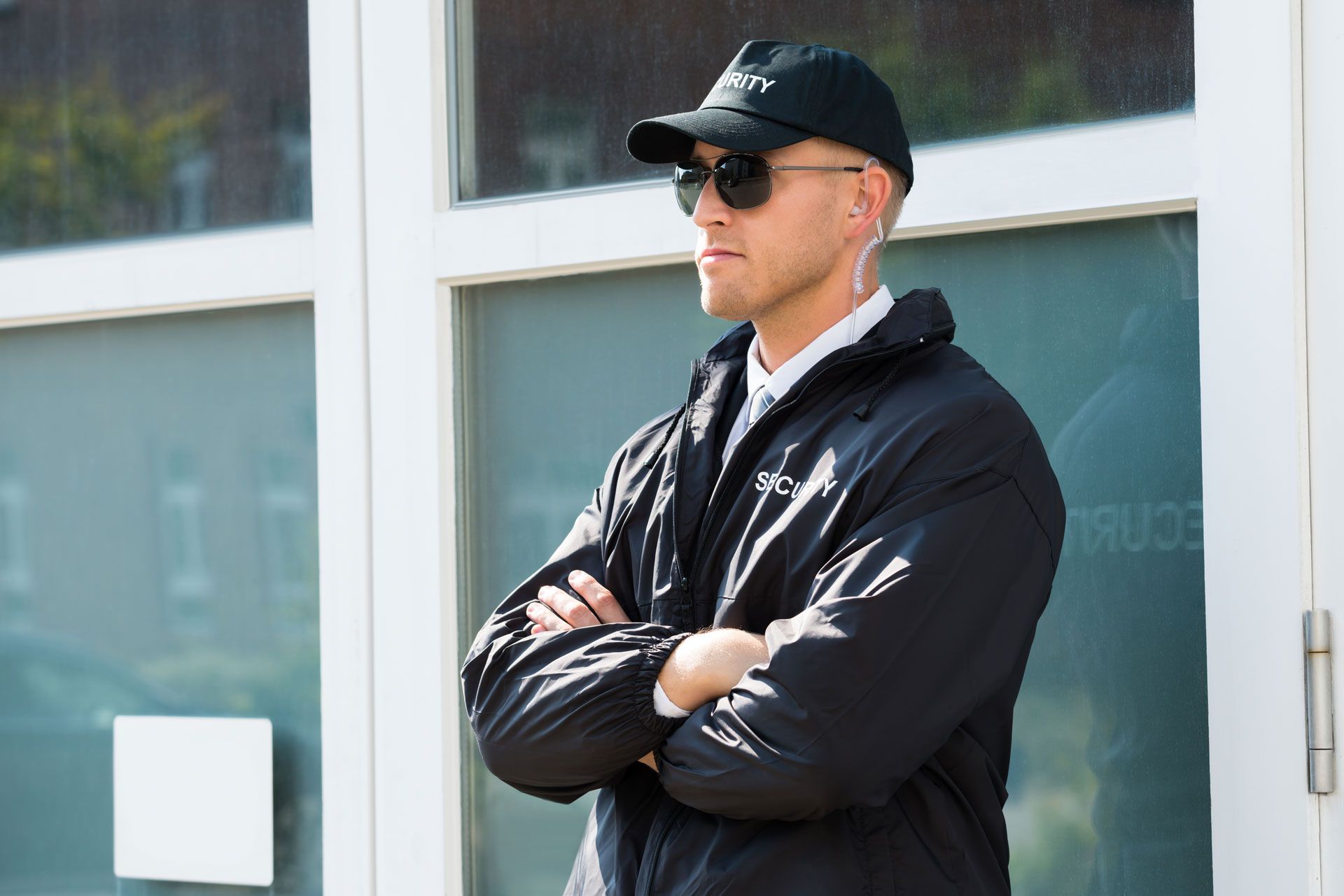 Security guard, black jacket, sunglasses, cap, arms crossed, standing in front of glass doors.