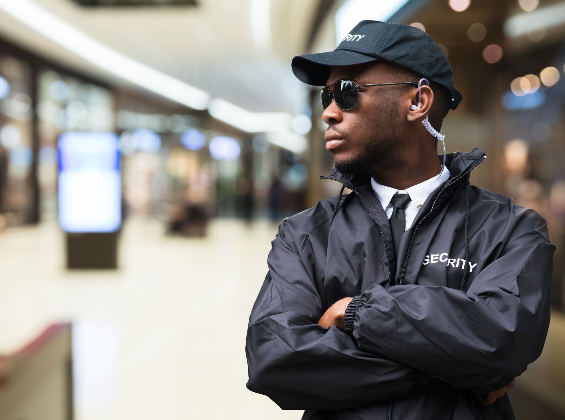 Security guard in black uniform with crossed arms, sunglasses, and earpiece in a shopping mall.