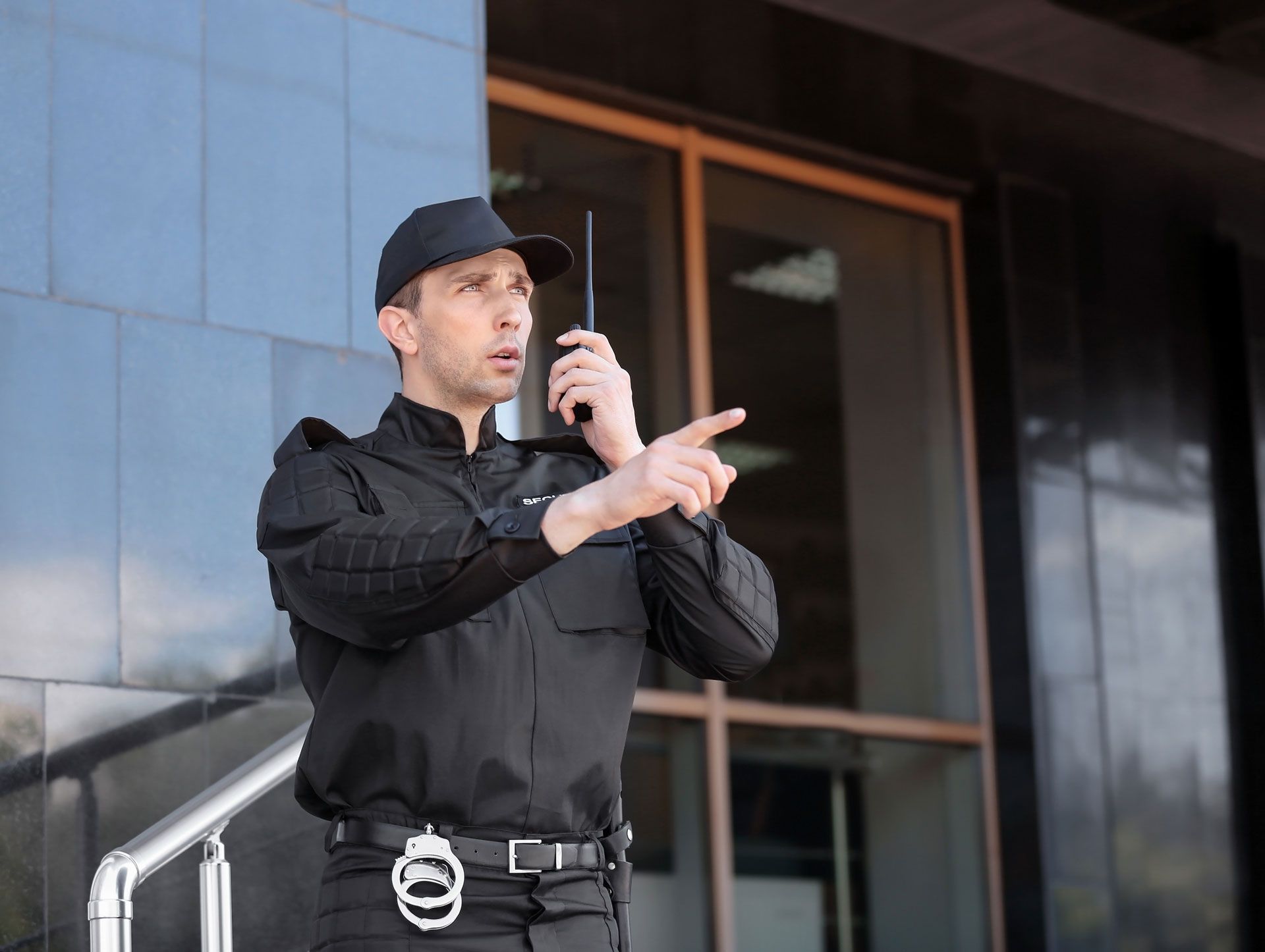 Security guard in black uniform talking into a radio, pointing to the right, near a building entrance.