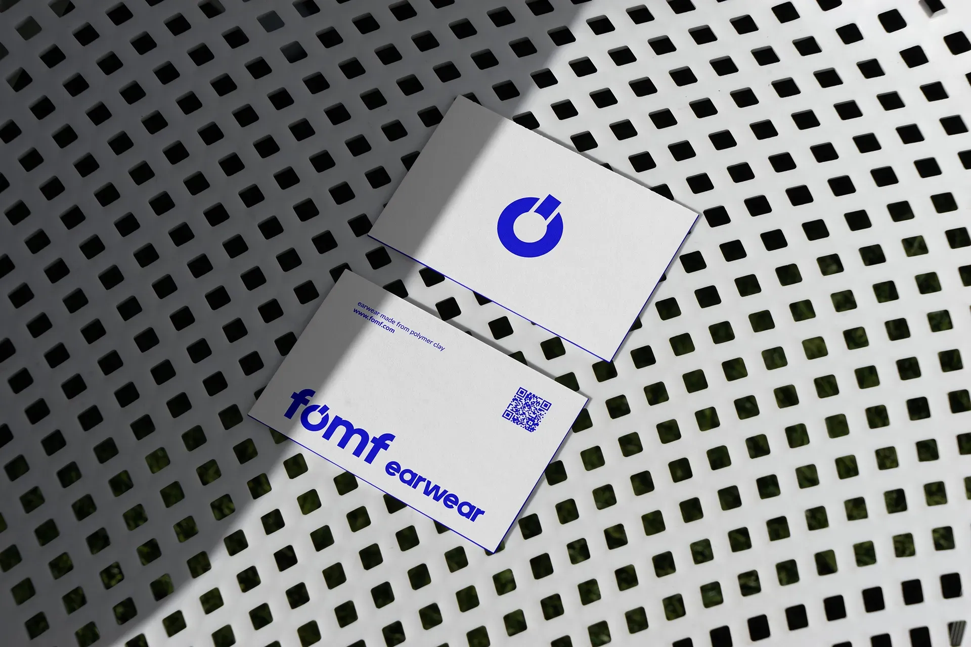 Zwei weiße Visitenkarten auf einer perforierten Metalloberfläche, eine mit einem blauen Logo und eine mit der Aufschrift „fomf earwear“.