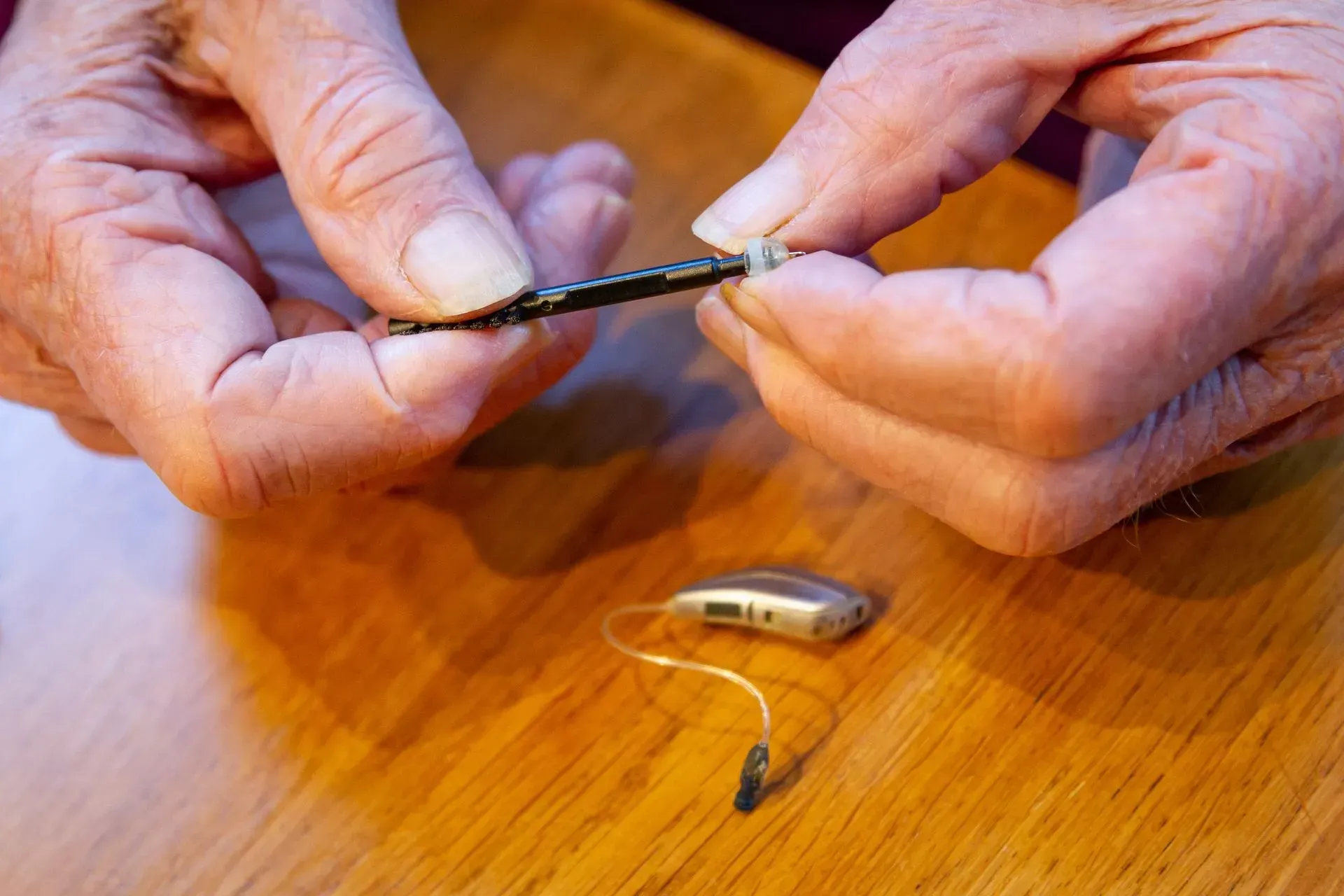 Hands repairing a small hearing aid on a wooden table.