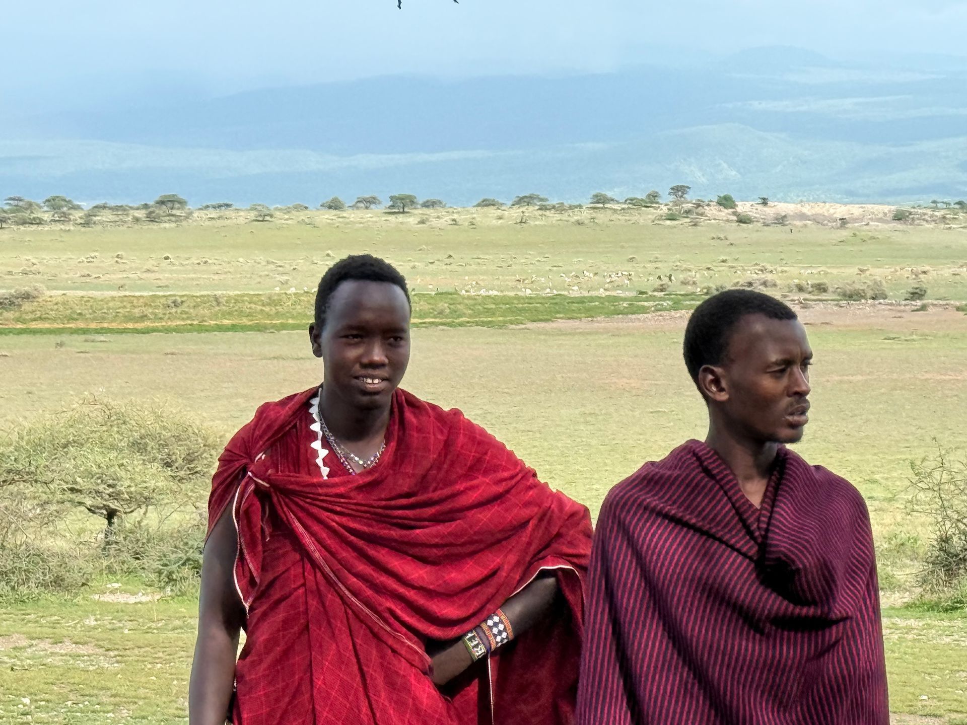 Two Maasai men in traditional robes, standing in a grassy field, mountains in background.