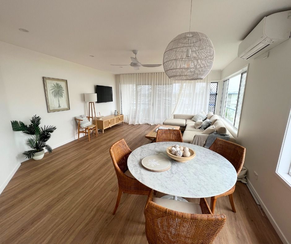 Living room with round dining table, couch, television, and large window. Wooden floors, light colors. — Nyblad Construction in Coolum, QLD