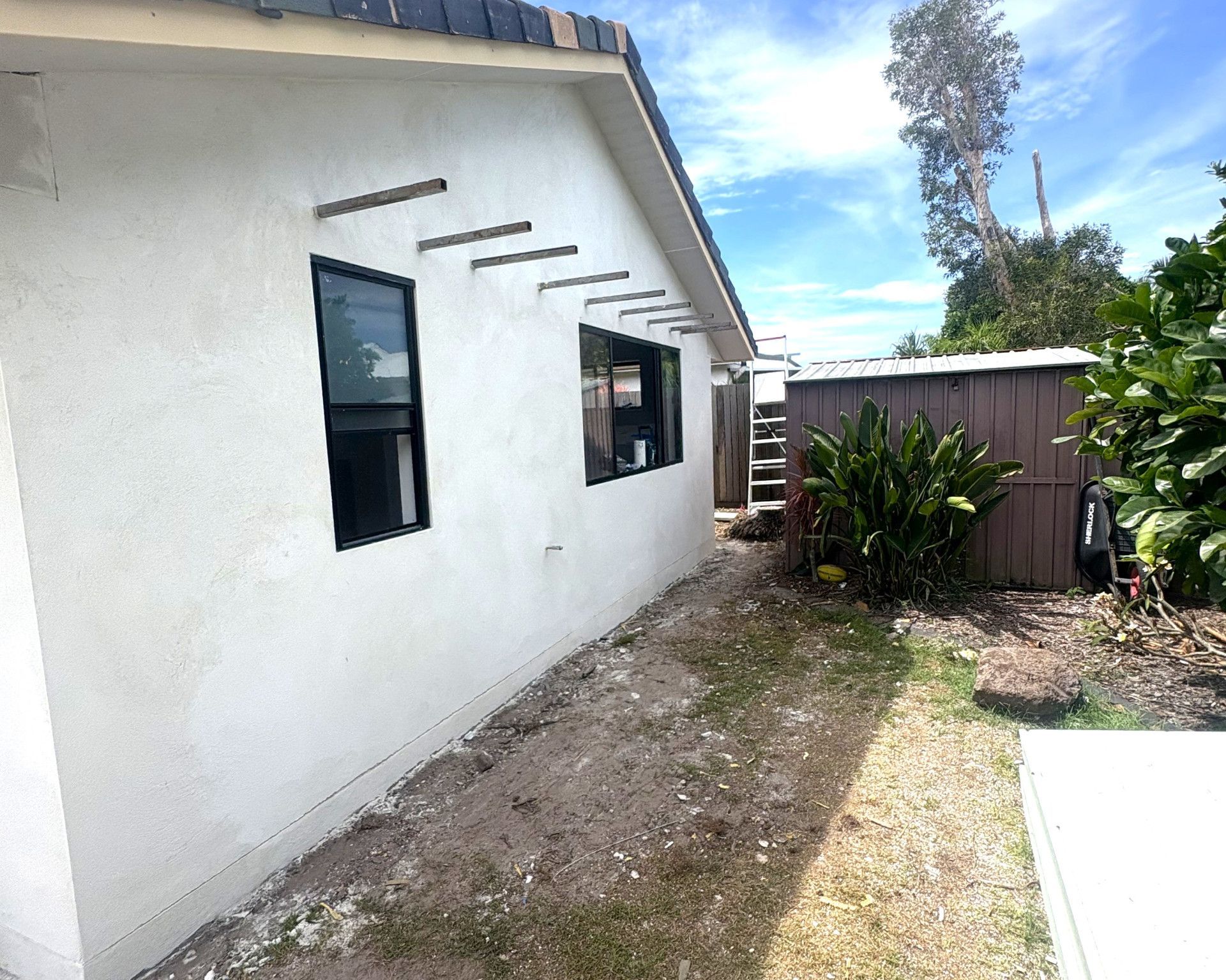White stucco house exterior with dark windows and wooden beams. Small shed and greenery on the side. — Nyblad Construction in Maroochydore, QLD