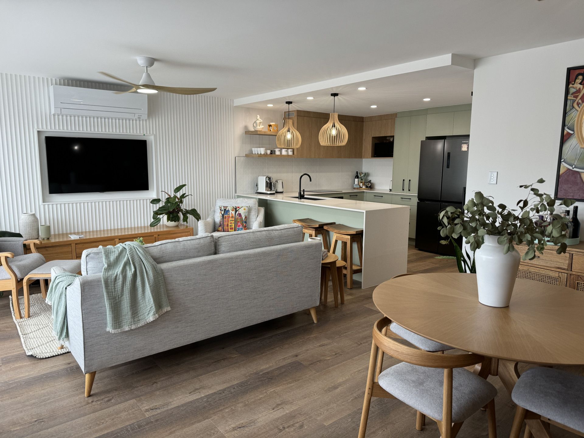 Modern open-plan living room with light grey sofa, wooden dining table, kitchen, and textured accent wall. — Nyblad Construction in Coolum, QLD
