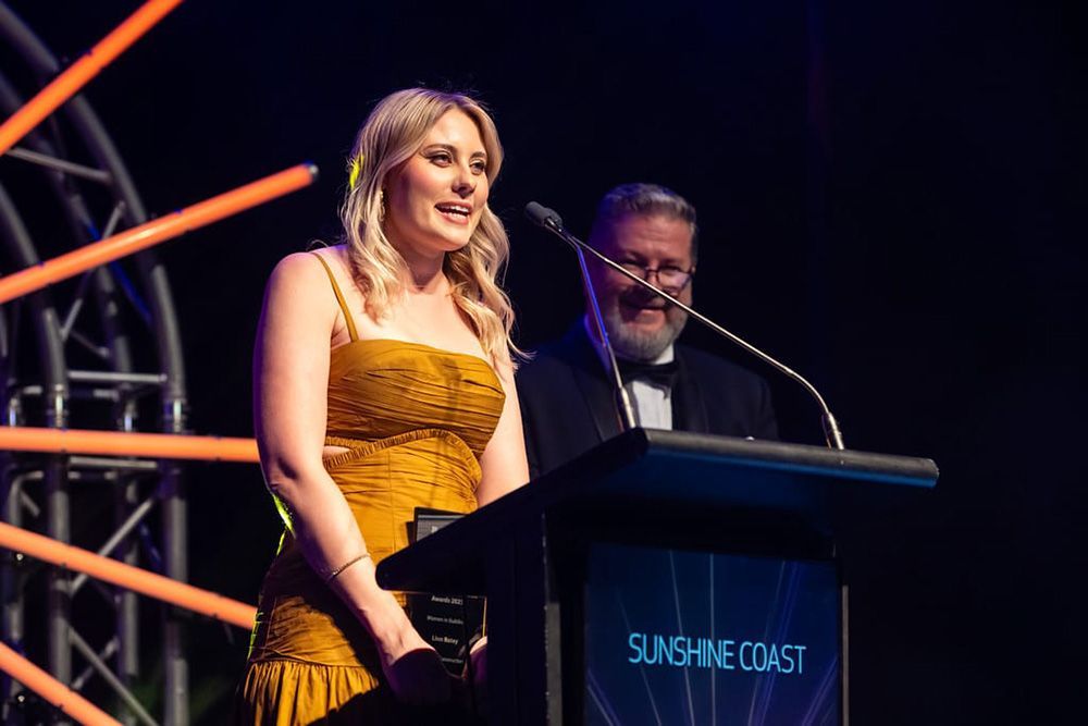 A Woman In A Yellow Dress Is Standing At A Podium — Nyblad Construction in Caloundra West, QLD
