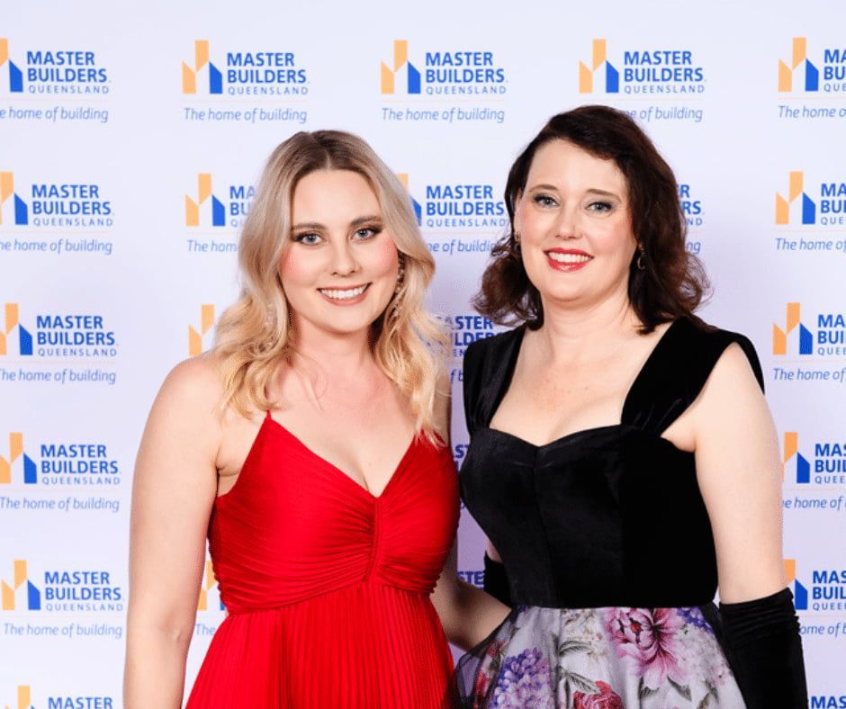 Two Women Are Posing for a Picture in Front of a Master Builders Wall — Nyblad Construction in Caloundra West, QLD