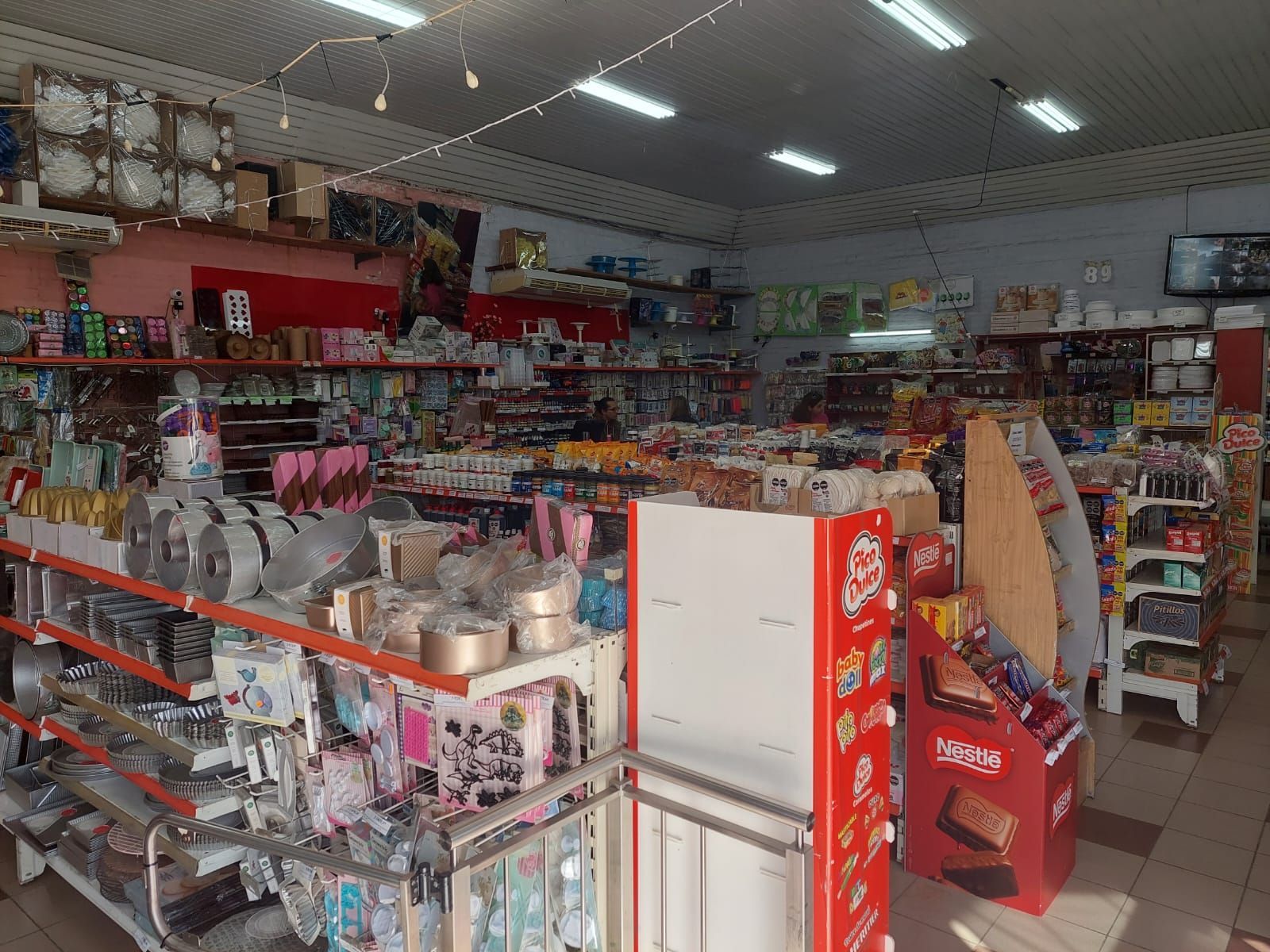 Pasillo de tienda de conveniencia con estantes de bocadillos, bebidas y artículos para el hogar bajo luces fluorescentes.