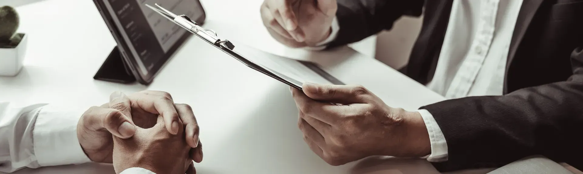 People in business attire in a meeting, hands gesturing over documents and a tablet on a white table