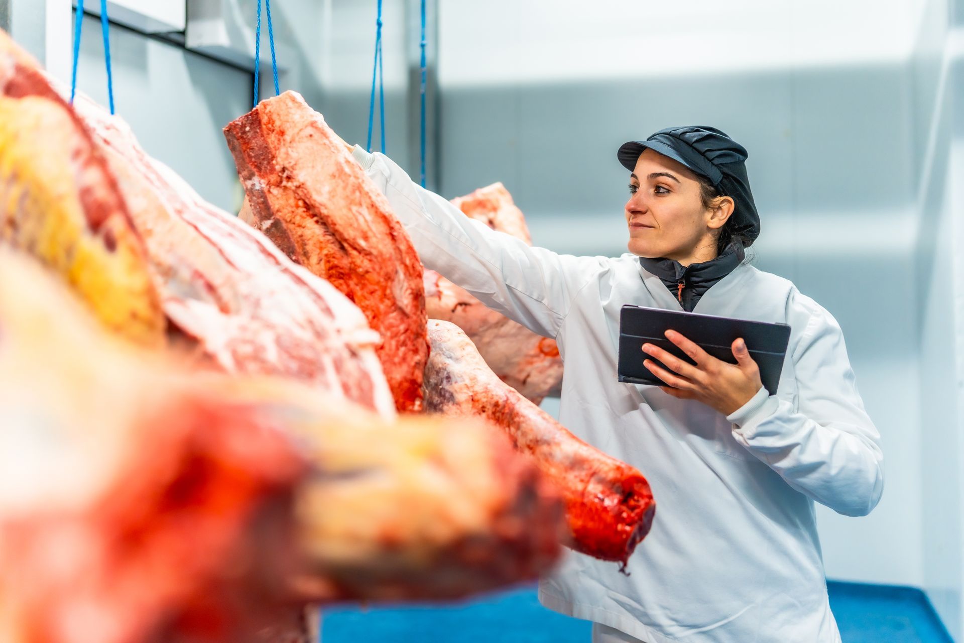 Worker inspecting hanging meat in a processing facility, holding a tablet