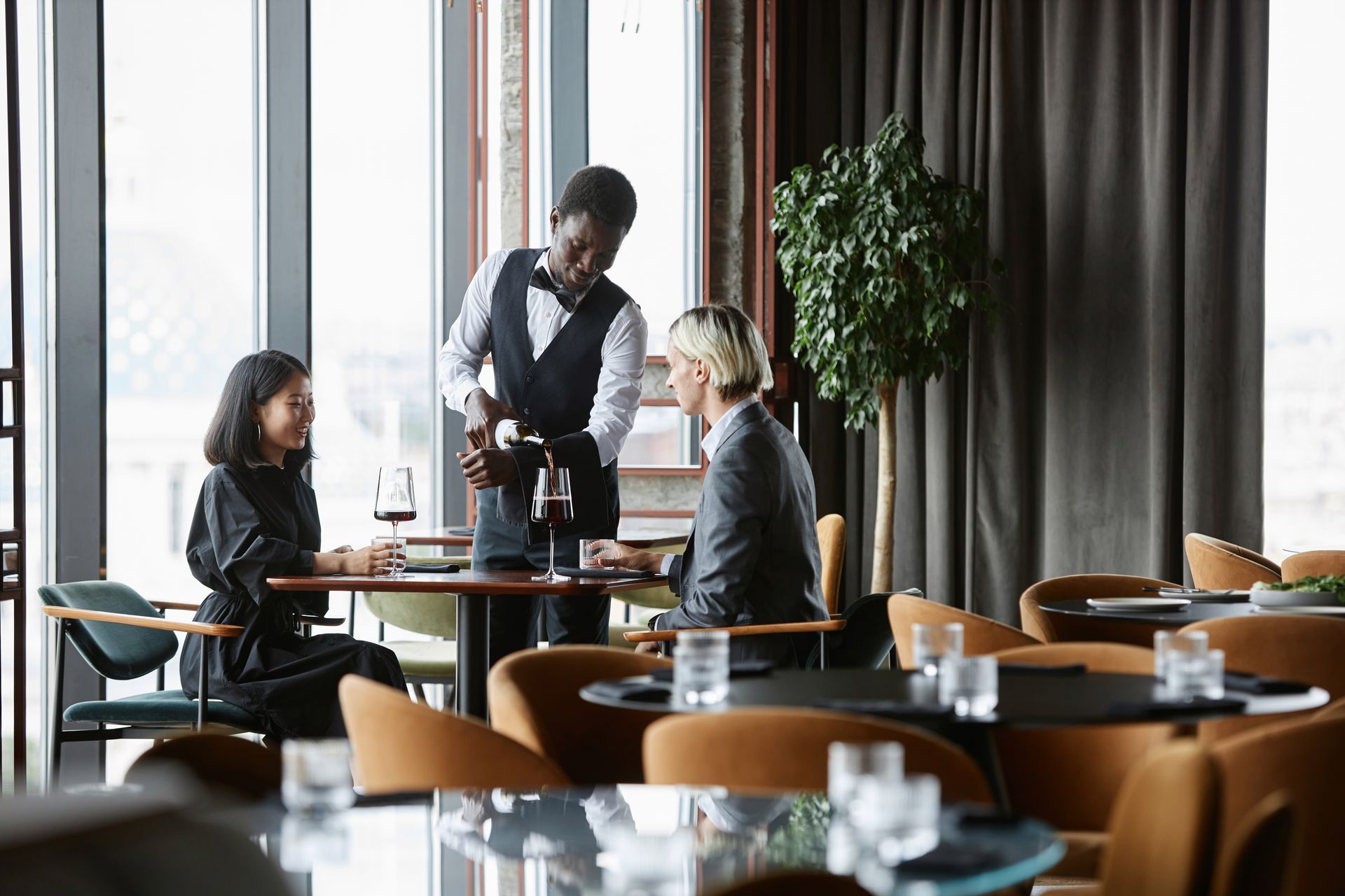 Waiter serving two diners in a modern restaurant with large windows and set tables