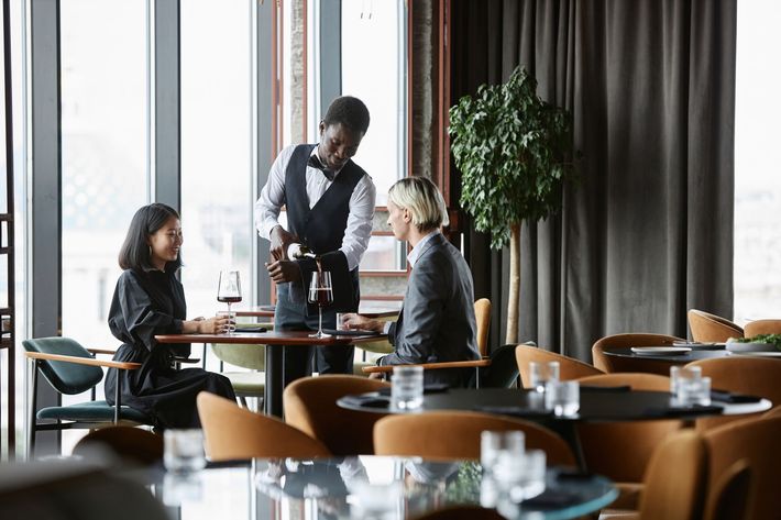Server taking an order at a restaurant table by a window, with two seated diners nearby.