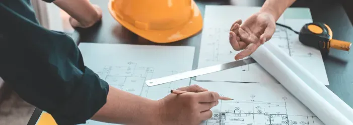 Hands reviewing architectural blueprints with a yellow hard hat and measuring tape on a desk