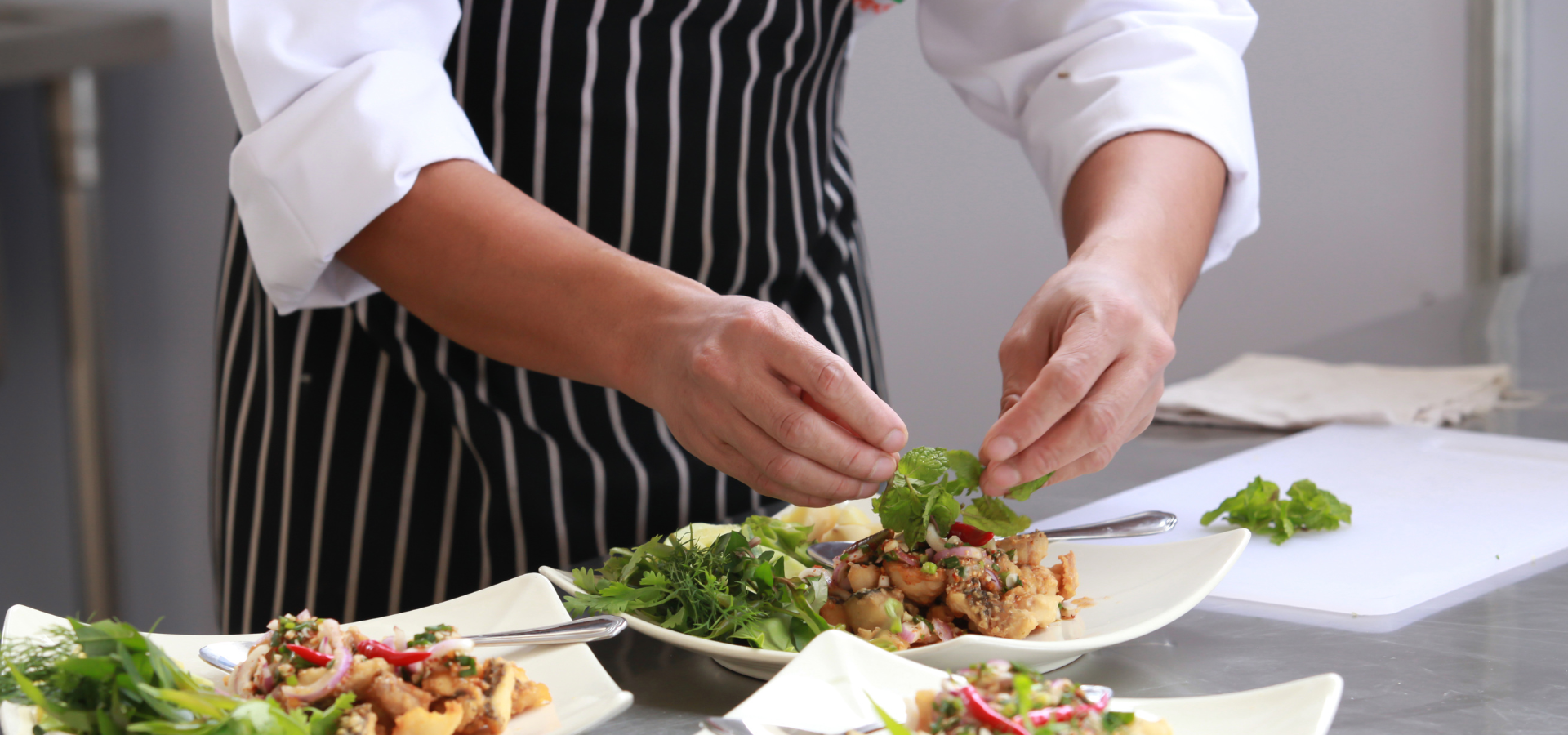 Chef plating a salad on a white table in a bright kitchen, wearing a striped apron