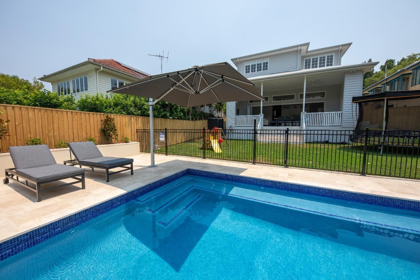 Backyard pool with lounge chairs, shaded patio, and a white house behind a black fence