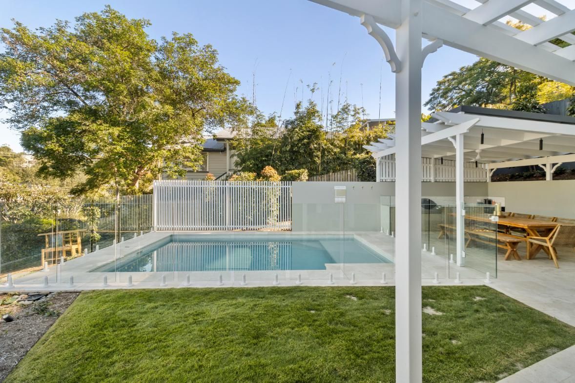 Backyard swimming pool beside a white pergola, patio seating, and green lawn at sunset