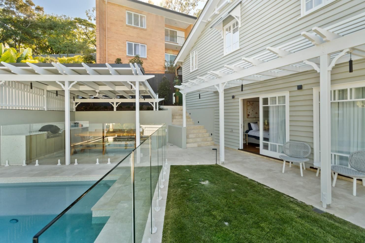 Backyard patio with swimming pool, white pergola, glass fence, and brick house facade