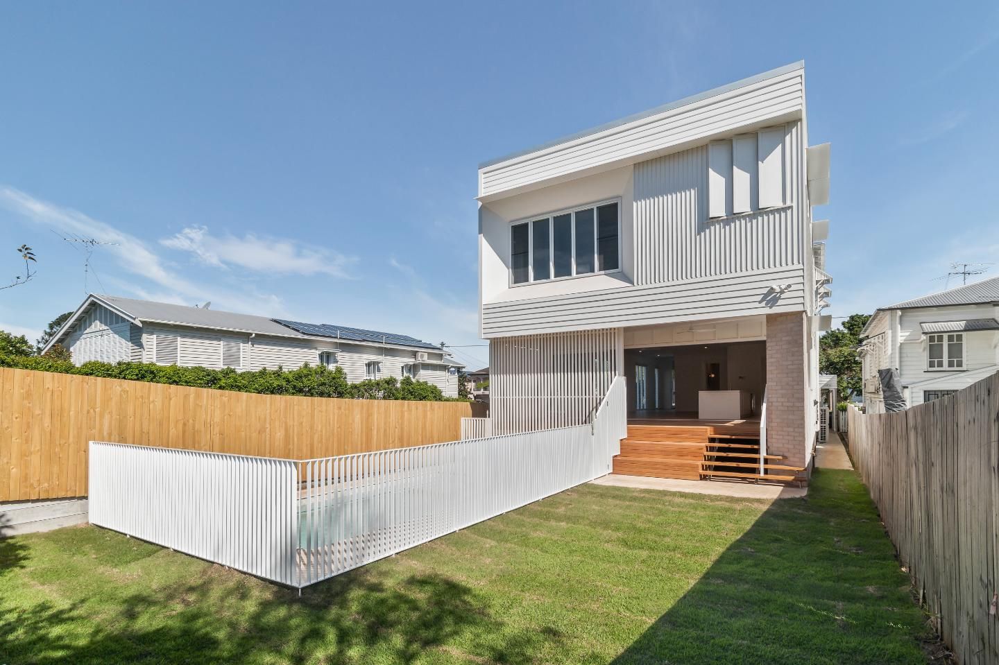 Modern two-story house with white fencing, wooden front porch, and green lawn under a blue sky