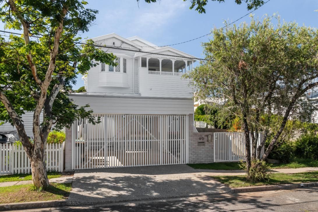 White two-story house with a gated front porch, trees, and a picket fence along a quiet street
