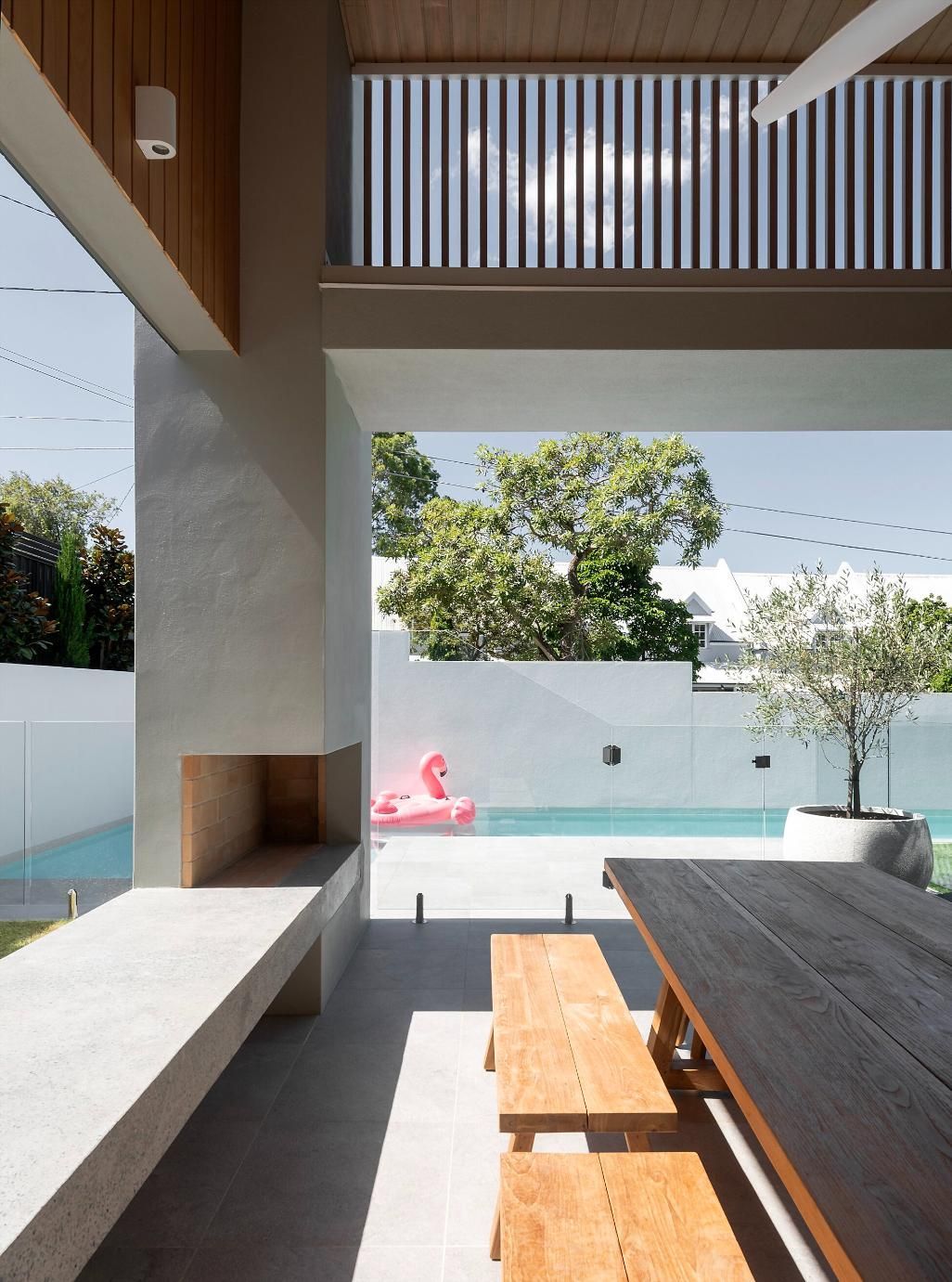 Modern patio with a long dining table, wooden benches, concrete fireplace, and poolside view.