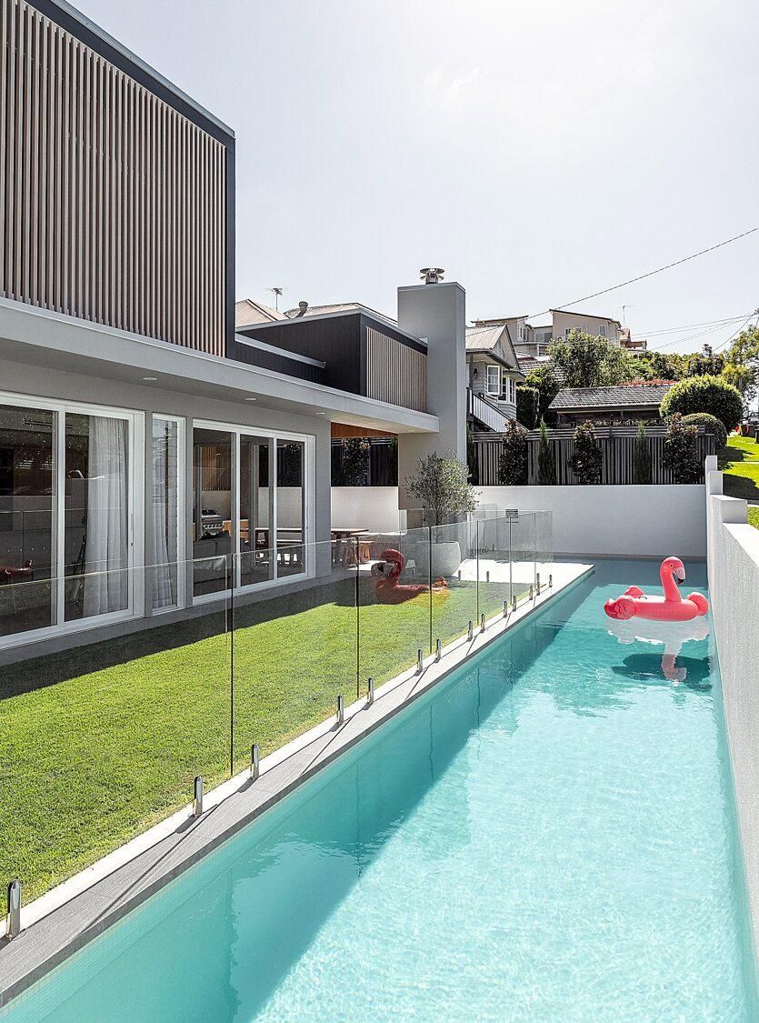 Modern backyard with a long turquoise pool beside a glass-walled house and lawn, with a pink float.