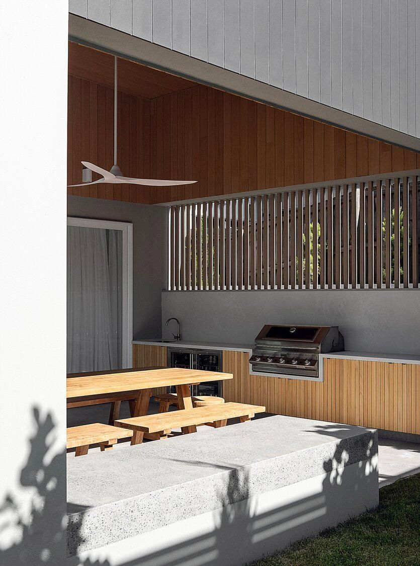 Modern patio with wooden picnic tables, built-in grill, and a white cushioned bench under a slatted roof