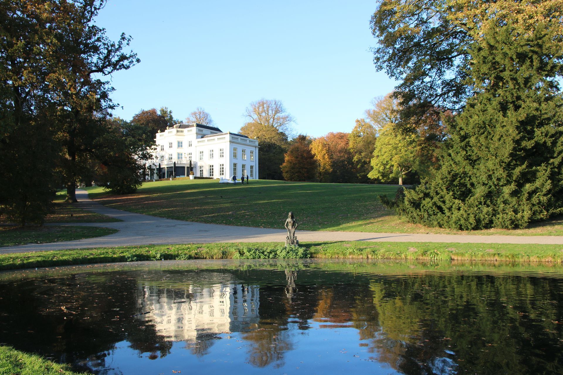 Huis Sonsbeek. Een wit landhuis naast een vijver, met bomen en herfstbladeren die in het water weerspiegeld worden.
