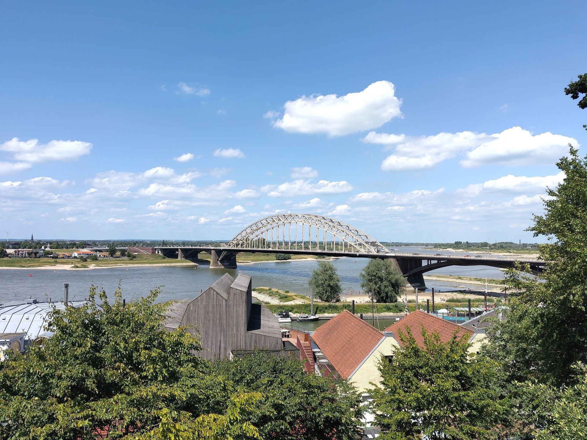 De WaalbrugEen brede rivier met een brug en in de verte een boogbrug onder een blauwe hemel met wolken, daken en bomen op de voorgrond.