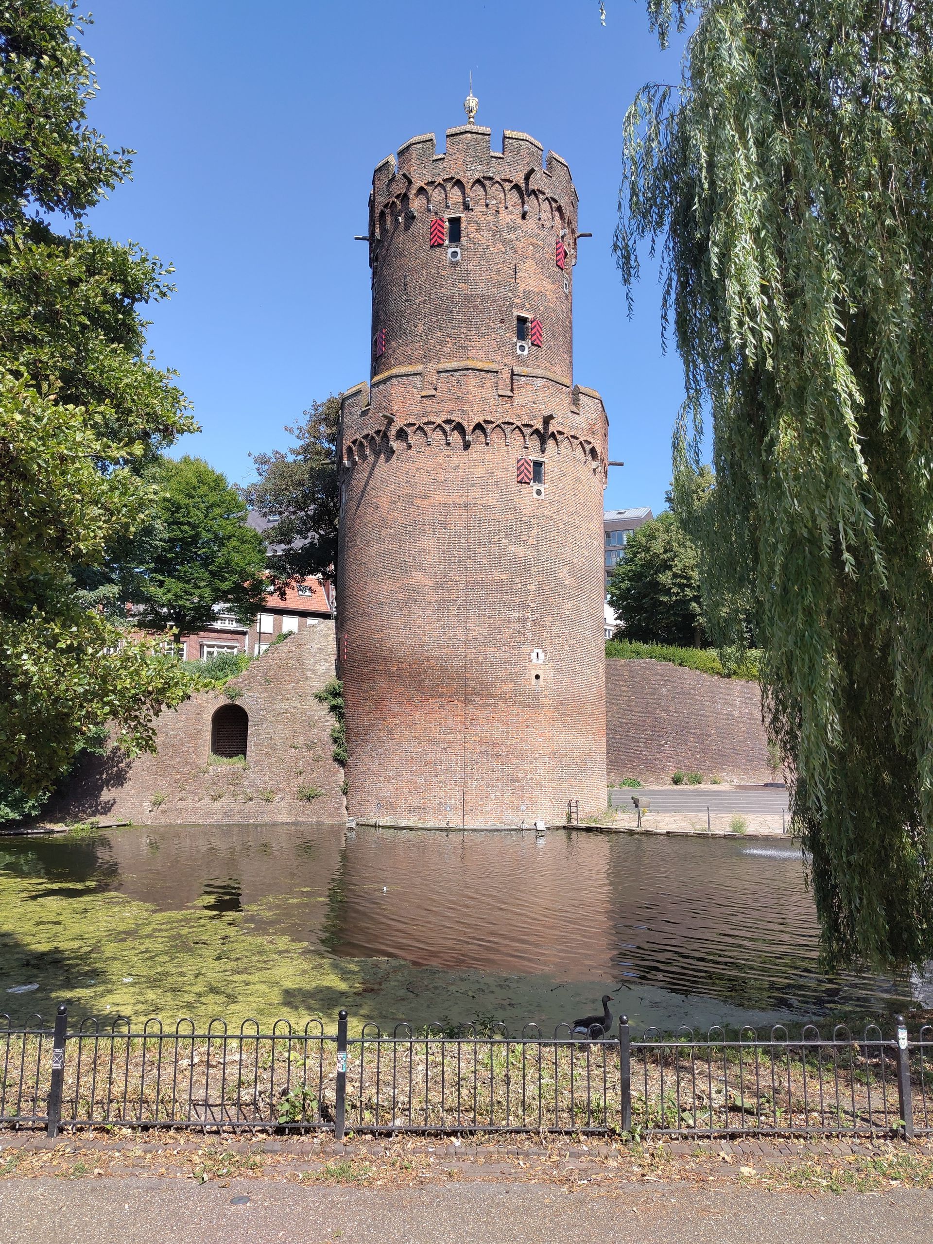 Ronde bakstenen kasteeltoren aan een gracht, omgeven door bomen onder een helderblauwe hemel.