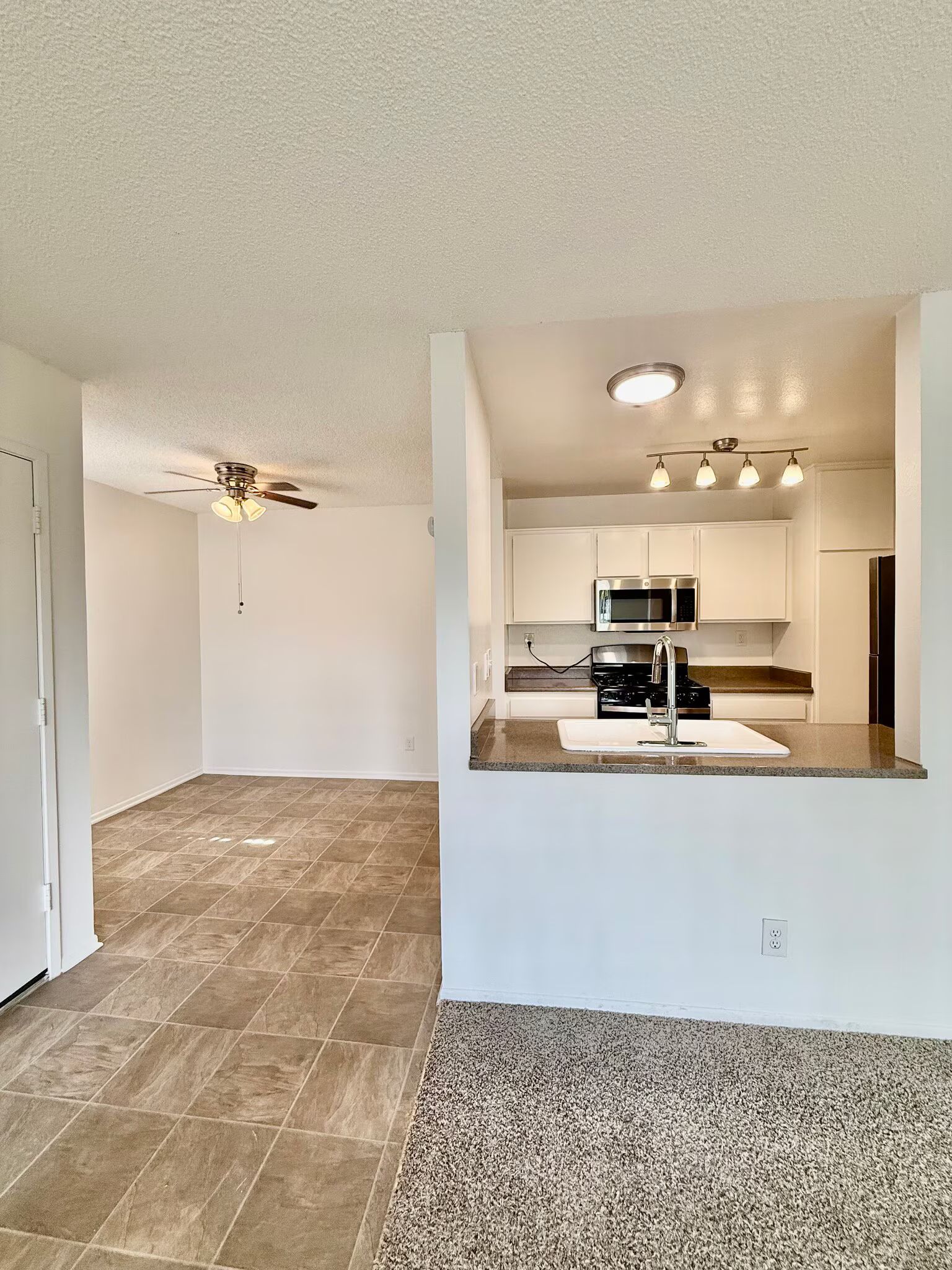 Empty open-plan living area with tile and carpet, looking into a kitchen with bar counter and ceiling lights