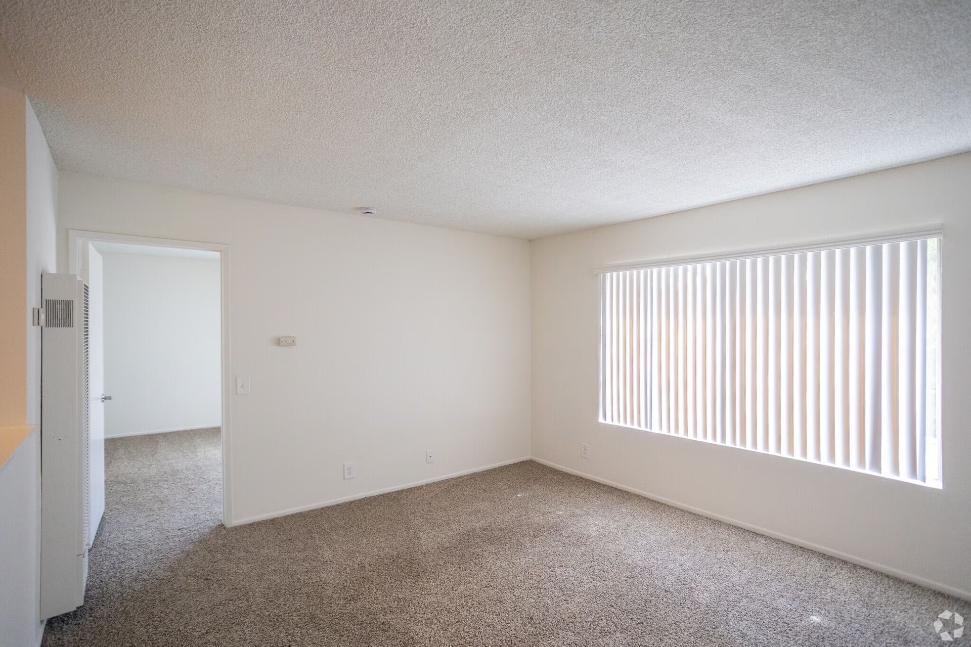 Empty carpeted room with white walls, doorway, and a large window with vertical blinds.