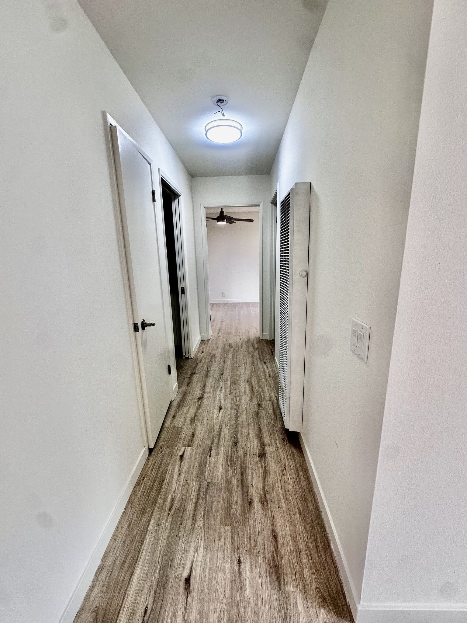 Narrow white hallway with wood-look floor and ceiling light, viewed toward a doorway at the end