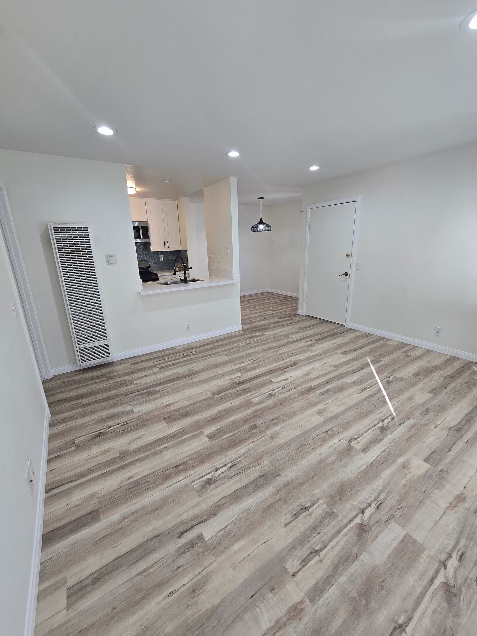 Empty open-plan room with wood-look flooring, white walls, recessed lights, and a small kitchen pass-through.